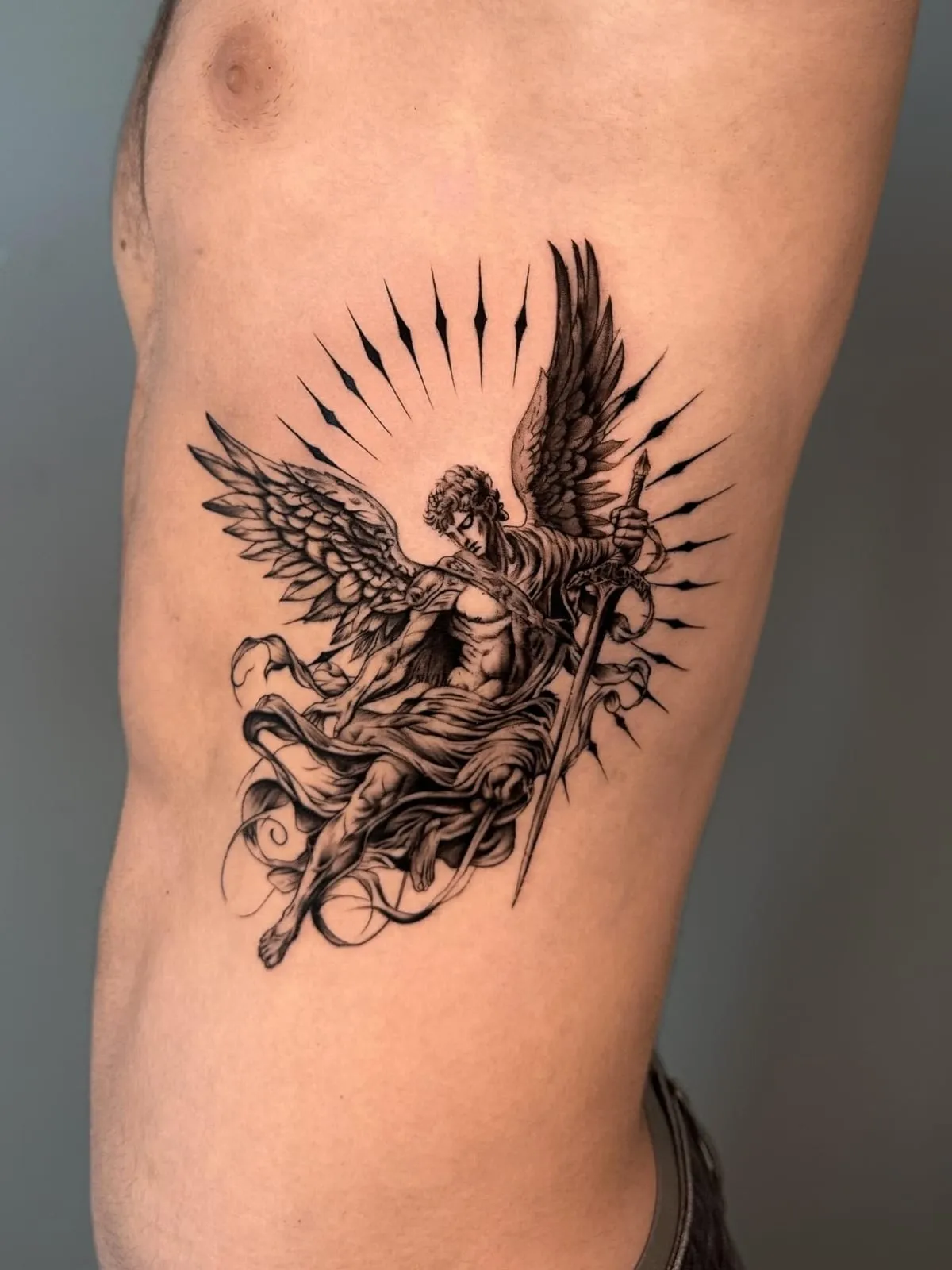 Black and grey realism tattoo of an archangel with spread wings holding a sword, surrounded by rays, on the ribs.