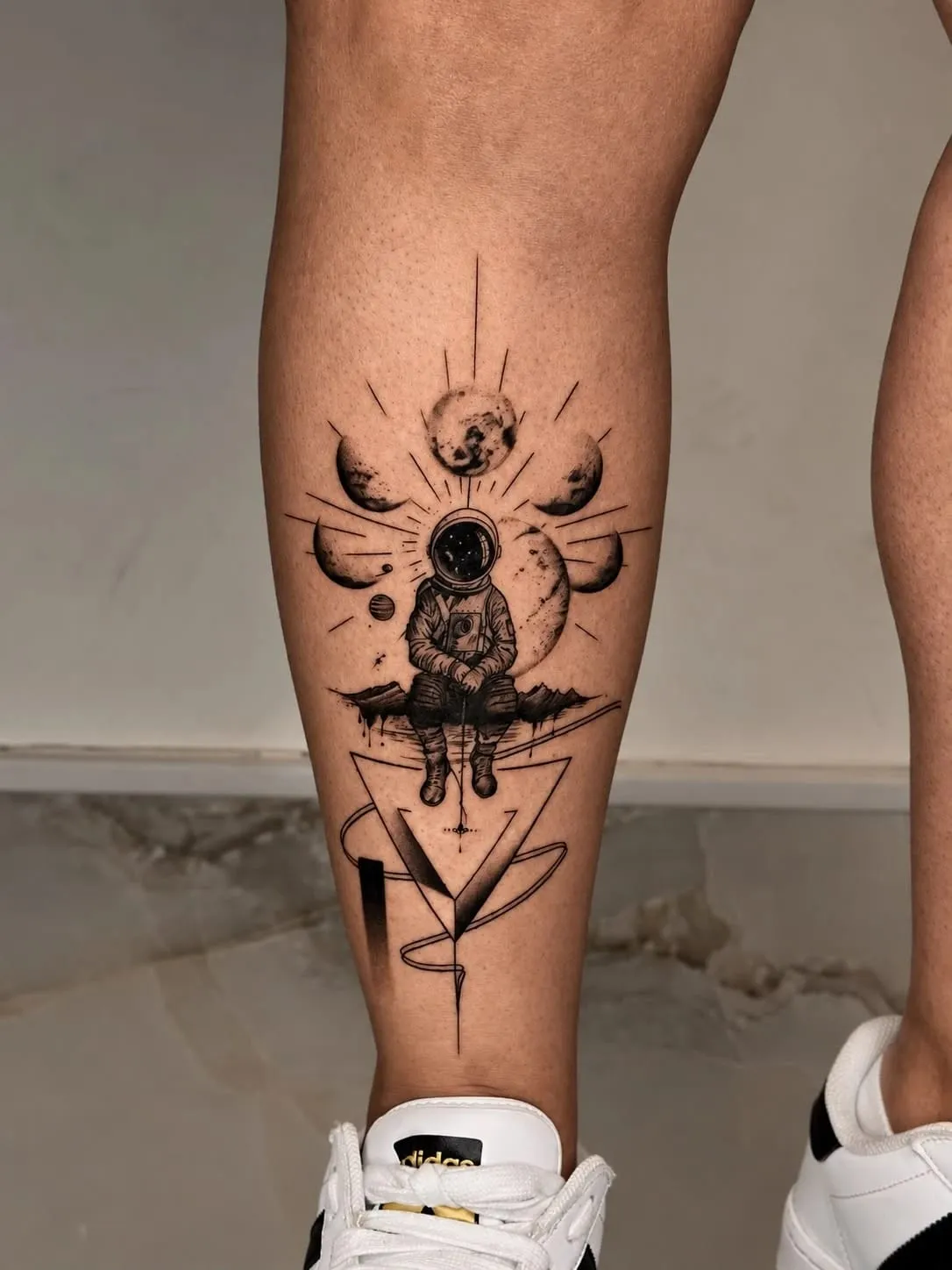 Astronaut sitting on a mountain under a row of moon phases and rays, with a geometric triangle below, on the calf.