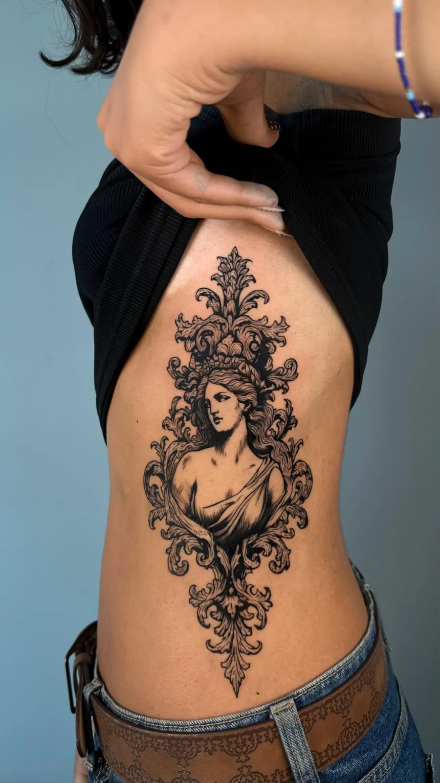 Engraving-style tattoo of a woman's bust inside an ornate baroque frame, on the ribs.