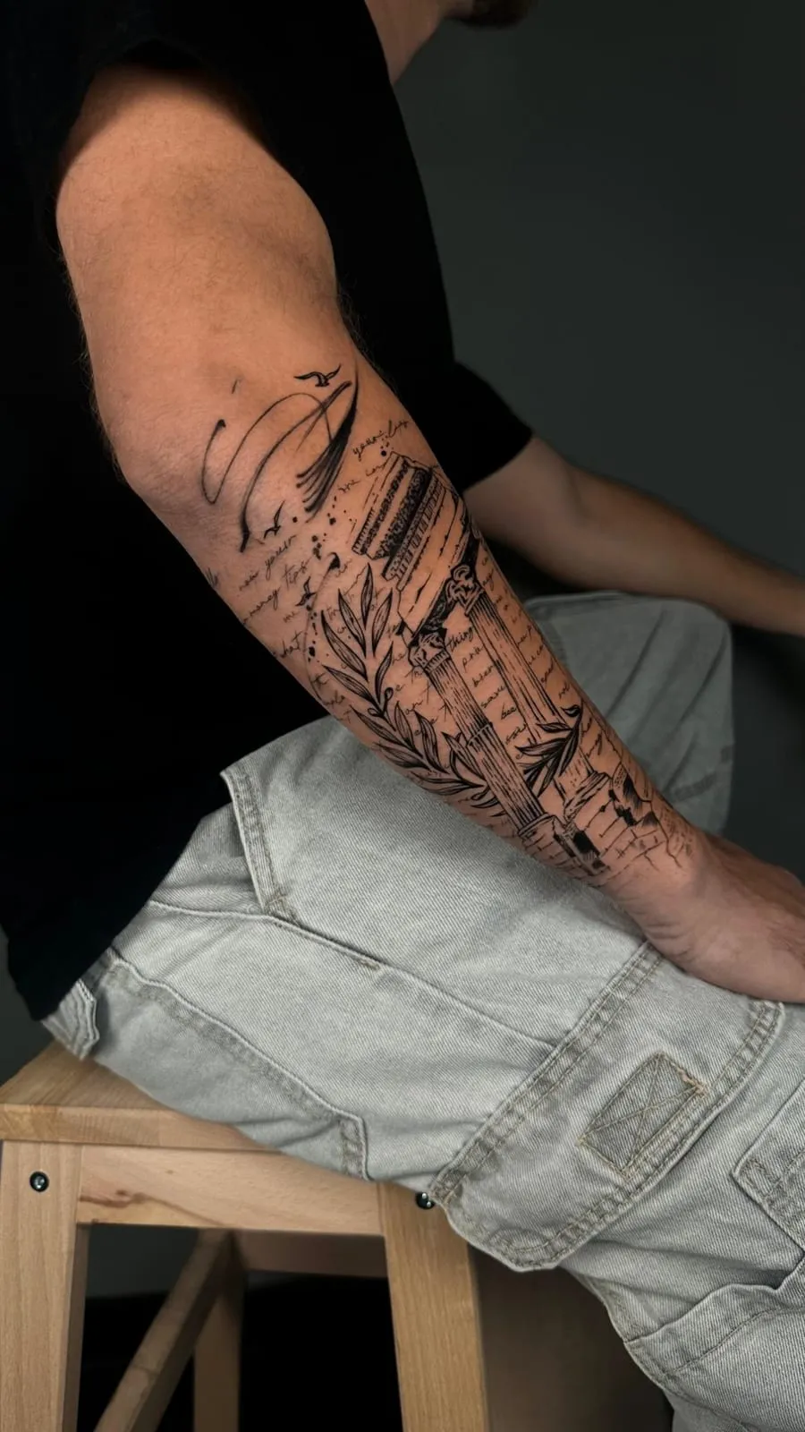 Fine-line ruined classical column with a laurel branch, birds and handwriting, on the forearm.