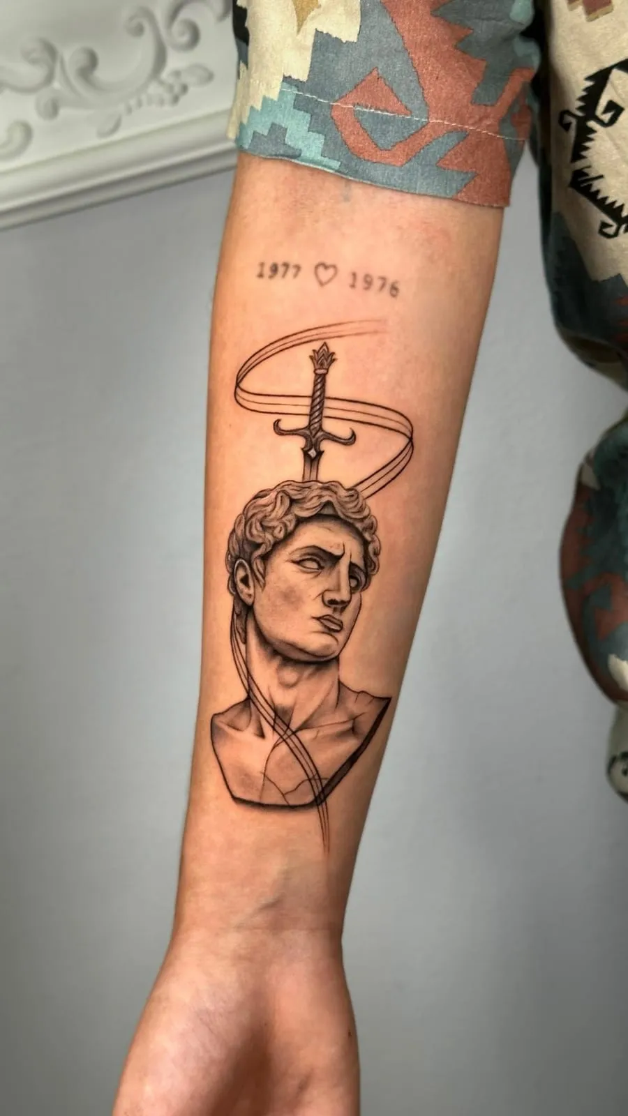 Black and grey tattoo of the bust of Michelangelo's David with a sword and a ribbon above it, on the forearm.