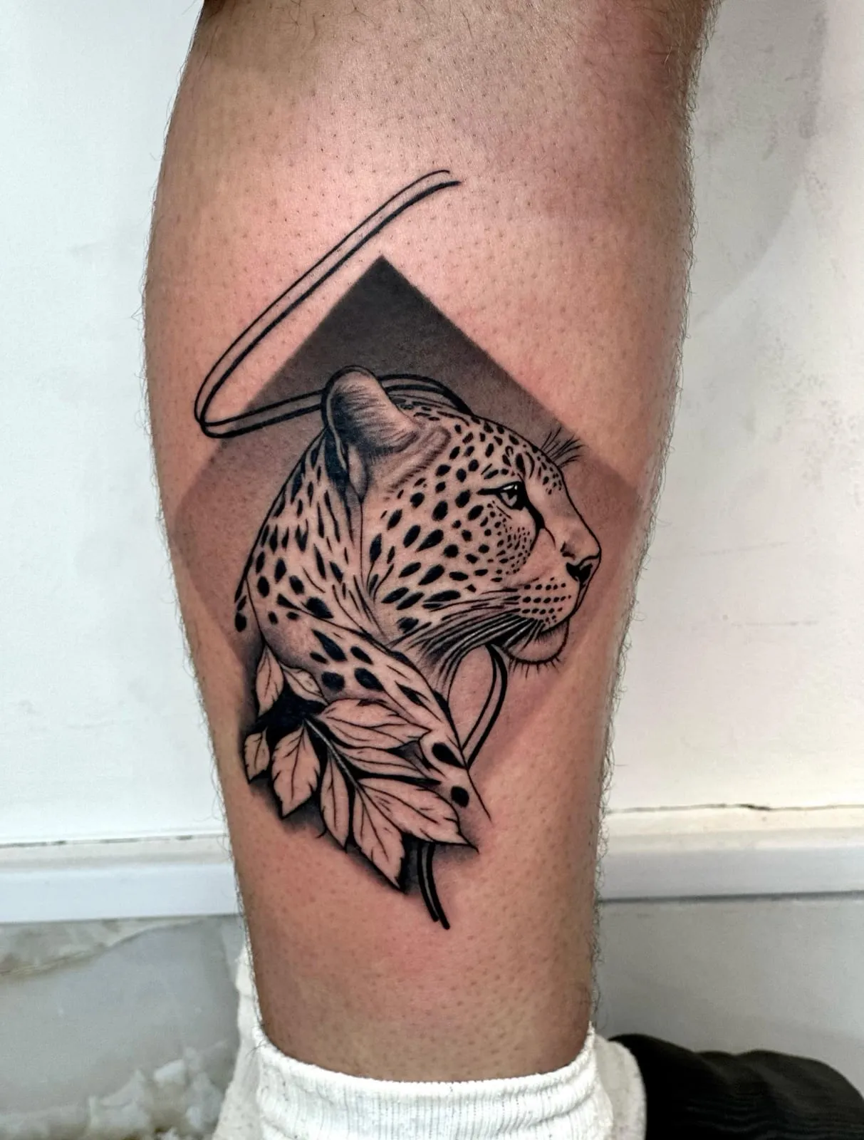 Black and grey realism leopard head with leaves, set against a shaded diamond and a fine arc, on the calf.