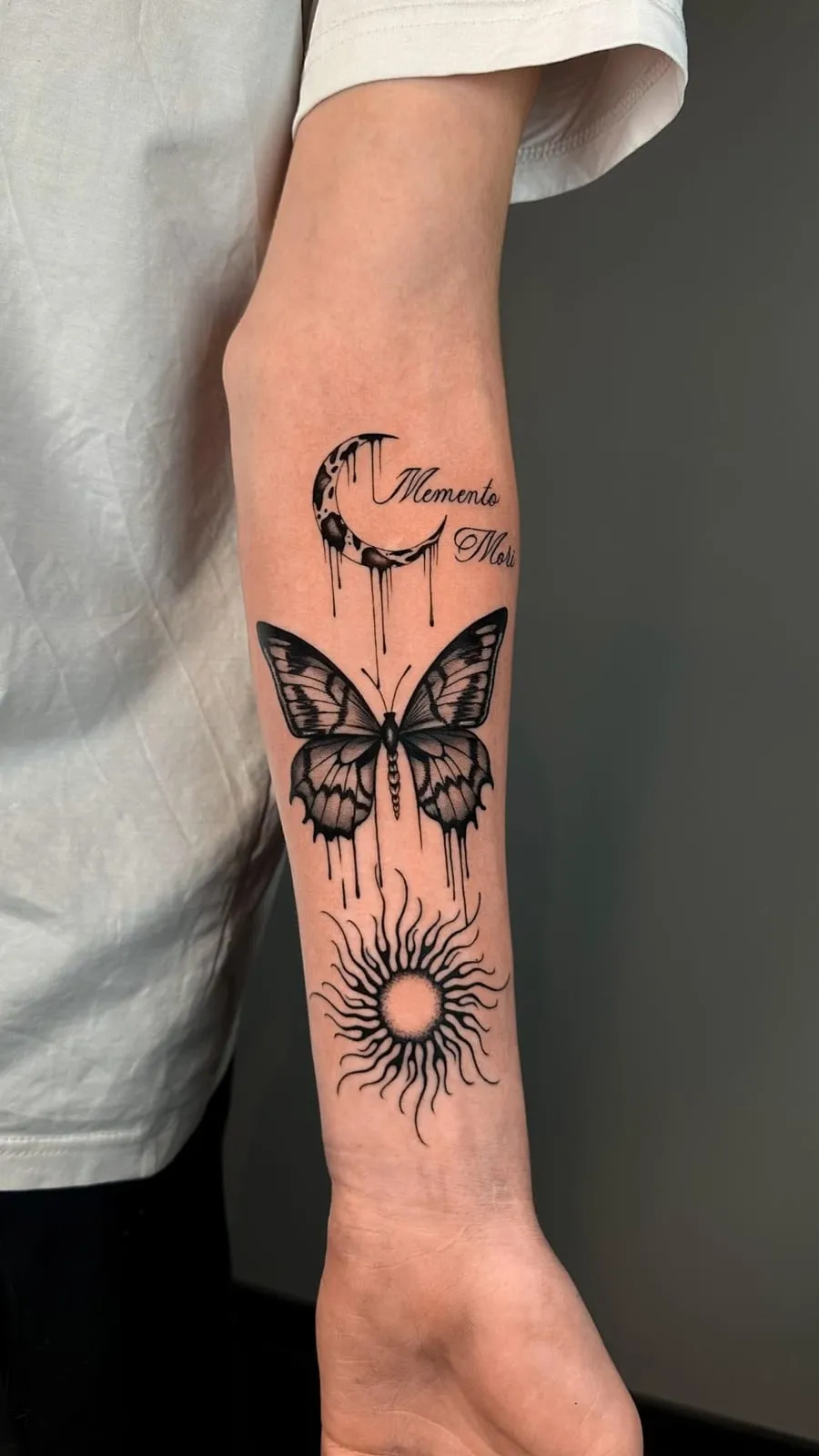 Dripping crescent moon with the words Memento Mori, a butterfly and a sun, on the forearm.