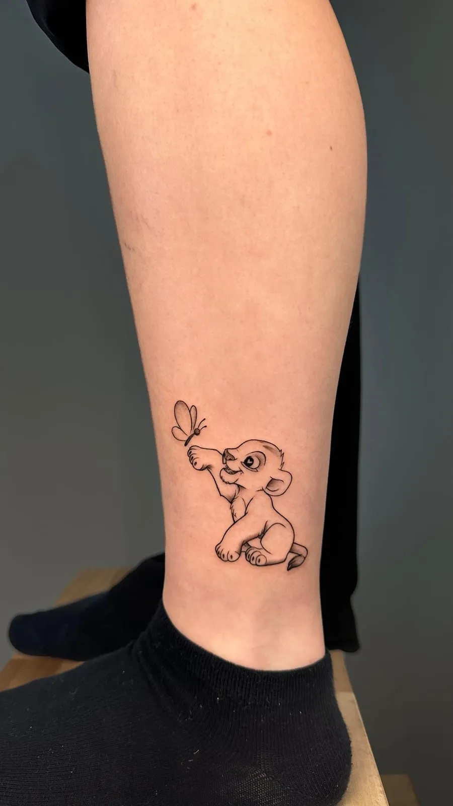 Young Simba reaching for a butterfly, in fine line, on the lower leg.
