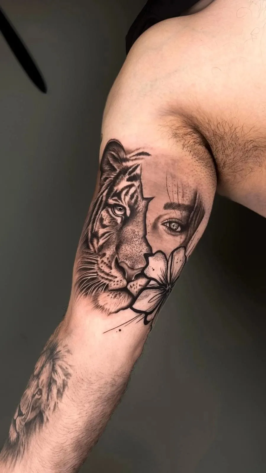 Realism tattoo of a face split between a tiger and a woman, with a fine-line flower, on the inner upper arm.